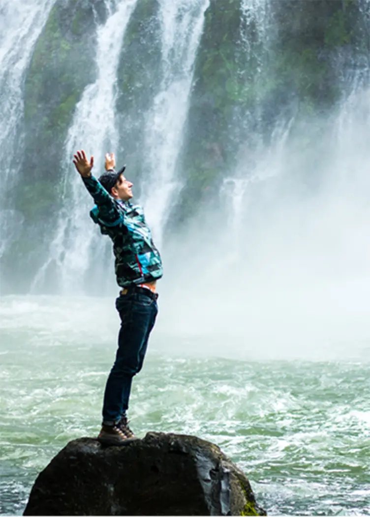 A man on a rock reaching into the sky in front of a waterfall